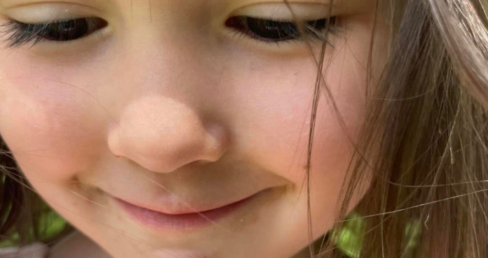extreme closeup of a young girl's face, slightly smiling as she looks down at something below outside of the photo. her brunette hair wisps around her face. there is a green background behind her mostly obscured, indicating an outdoor location.