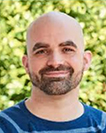 Image of presenter Nate Sheets; white male, bald with dark brown beard, wearing a blue shirt, he is smiling and the image shows him from the neck up, with blurred image of green leafy trees or bushes behind him.
